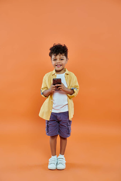 niño preescolar afroamericano feliz en traje casual usando teléfono inteligente sobre fondo naranja - Foto, imagen