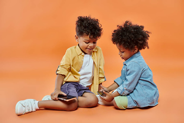 smiling african american boy in casual attire looking at toddler brother playing on smartphone - Zdjęcie, obraz