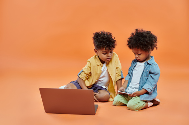 ノートパソコンの近くでスマートフォンで遊んでいるトッドラーの兄を見てカジュアルな服装をしているアフリカ系アメリカ人の少年 - 写真・画像