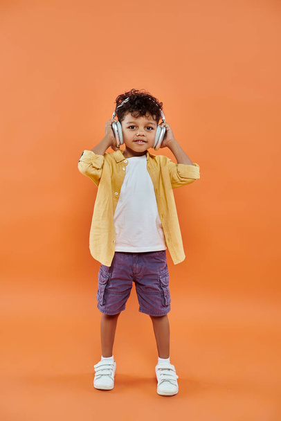 veselý a kudrnatý africký americký chlapec poslech hudby v bezdrátových sluchátkách na oranžovém pozadí - Fotografie, Obrázek