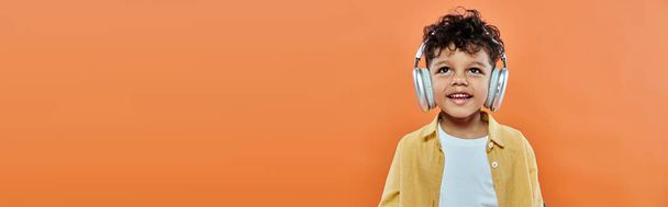 niño afroamericano feliz y rizado escuchando música en auriculares inalámbricos en naranja, bandera - Foto, Imagen