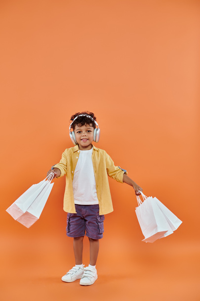 陽気なアフリカ系アメリカ人の少年がヘッドフォンで音楽を聴き,オレンジ色のショッピングバッグを持っている - 写真・画像