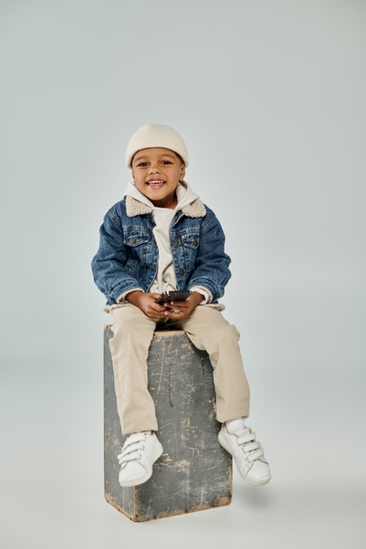 happy african american boy in winter attire and beanie using smartphone and sitting on concrete cube - Fotó, kép