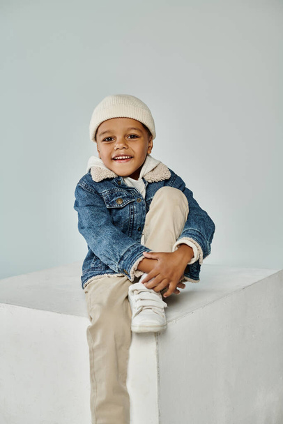 happy african american boy in winter attire and beanie hat sitting on concrete cube on grey - Photo, Image