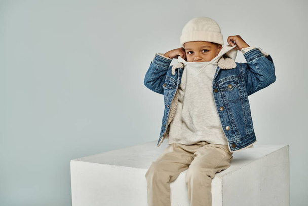 cute african american boy in winter attire and beanie sitting on concrete cube and adjusting hood - Photo, Image