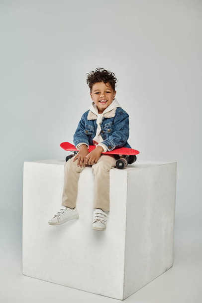 χαρούμενο αφροαμερικανό αγόρι σε χειμερινή ενδυμασία κάθεται σε κύβο με penny board σε γκρι φόντο - Φωτογραφία, εικόνα