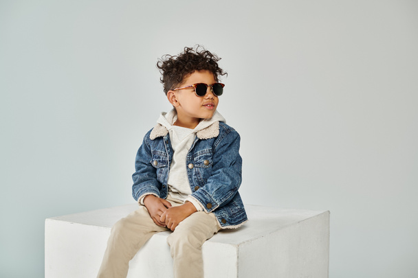 roztomilý a kudrnatý africký americký chlapec v zimě oblečení a sluneční brýle sedí na kostce na šedém pozadí - Fotografie, Obrázek