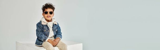 ricci ragazzo africano americano in abbigliamento invernale e occhiali da sole seduti sul cubo su sfondo grigio, banner - Foto, immagini