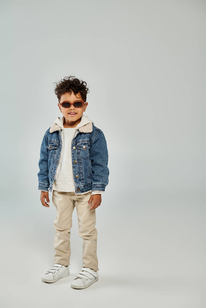 cheerful curly african american boy in winter attire and sunglasses standing on grey backdrop - Фото, изображение
