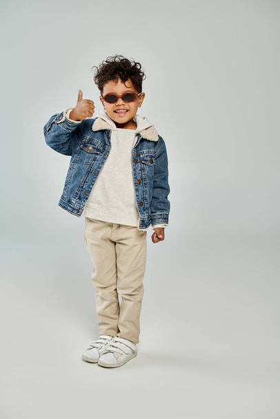 happy curly african american boy in winter attire and sunglasses showing thumb up on grey backdrop - Fotó, kép