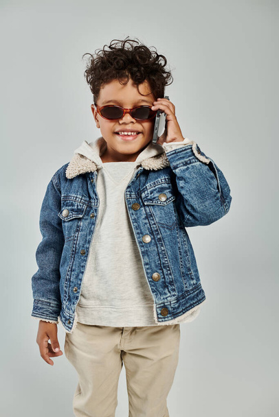 niño afroamericano feliz en traje de invierno y gafas de sol hablando en el teléfono inteligente sobre fondo gris - Foto, Imagen