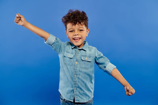 retrato de niño afroamericano emocionado en traje de mezclilla mirando a la cámara sobre fondo azul - Foto, Imagen