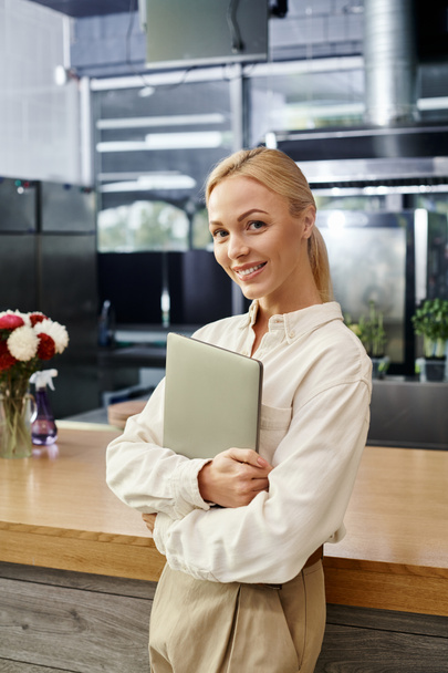 jolie administrateur de café blonde avec ordinateur portable souriant à la caméra près du comptoir, petite entreprise - Photo, image