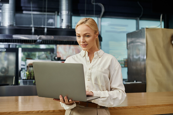 χαμογελαστή ξανθιά γυναίκα κοιτάζοντας φορητό υπολογιστή κοντά μετρητή στο σύγχρονο καφέ, επιχειρηματίας στην εργασία - Φωτογραφία, εικόνα