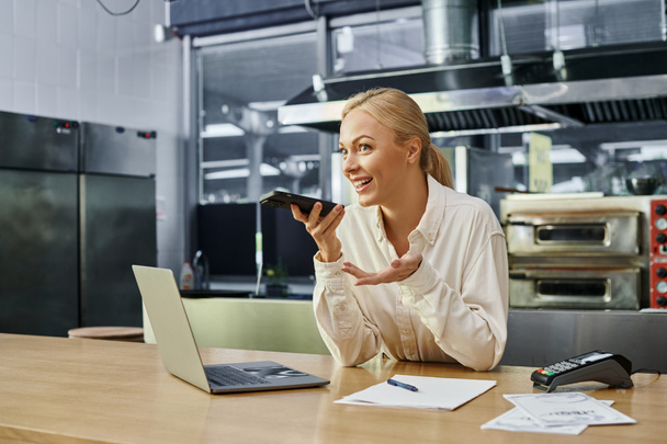 blondi johtaja lähettää ääniviestin älypuhelimella lähellä kannettavaa tietokonetta ja maksupäätteen kahvila laskuri - Valokuva, kuva