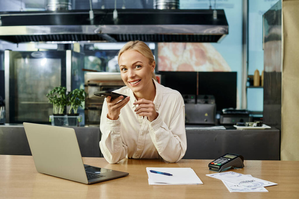 blondýny žena posílání hlasové zprávy na smartphone a usmívá se na kameru v blízkosti notebooku v kavárně - Fotografie, Obrázek