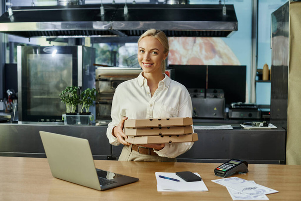 χαμογελαστή ξανθιά γυναίκα με κουτιά πίτσα κοντά σε ψηφιακές συσκευές και τερματικό πληρωμής στο cafe - Φωτογραφία, εικόνα