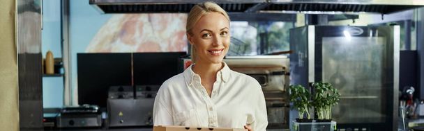 alegre rubia manager celebración pizza caja y sonriendo a la cámara en moderno café, pancarta horizontal - Foto, imagen