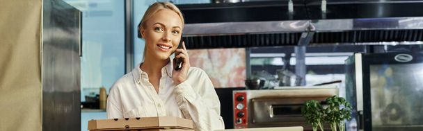 Lächelnde blonde Café-Managerin mit Pizzakartons, die auf dem Handy sprechen und in die Kamera schauen, Banner - Foto, Bild