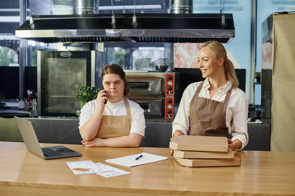 молодая женщина с синдромом Дауна разговаривает на смартфоне рядом с ноутбуком и кафе менеджер с коробками пиццы - Фото, изображение