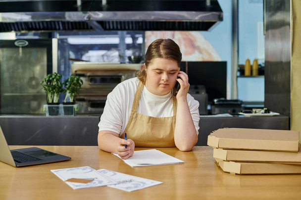 Down sendromlu genç kafe çalışanı bilgisayarın yanında akıllı telefondan konuşuyor ve tezgahta pizza kutuları var. - Fotoğraf, Görsel