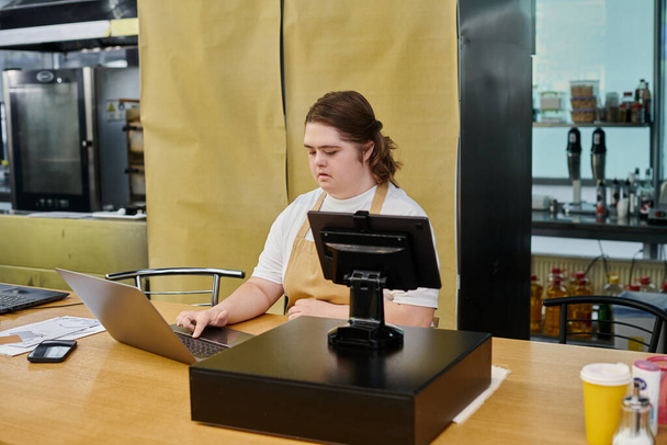 joven empleada con síndrome de Down que trabaja con portátil y terminal de efectivo en el mostrador en la cafetería - Foto, Imagen