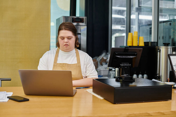 empleado femenino enfocado con discapacidad mental que trabaja en el ordenador portátil cerca de la terminal de efectivo en la cafetería moderna - Foto, imagen
