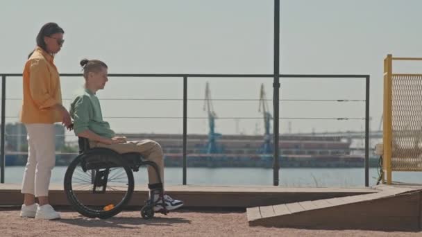 Genç bir kadın engelli bir adamın tahta tekerlekli sandalyeye binmesine yardım ediyor. Şehir köprüsünde asansörde. - Video, Çekim