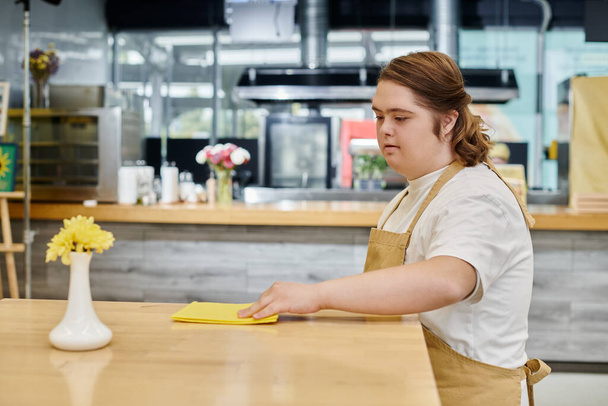młoda kobieta z zespołem Downa wycieranie tabeli z szmatą podczas pracy w nowoczesnej kawiarni, inkluzywność - Zdjęcie, obraz
