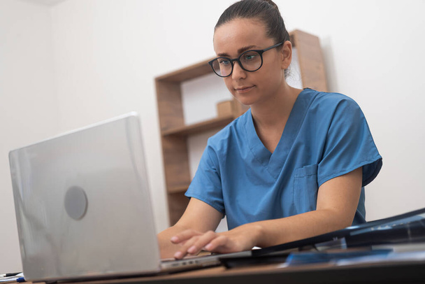 γιατρός με μπλε στολή χρησιμοποιεί το laptop της για να εξετάσει σχολαστικά τα αποτελέσματα των εξετάσεων των ασθενών κατά τη διάρκεια μιας online διαβούλευσης, επιδεικνύοντας την αποτελεσματικότητα της τηλεϊατρικής.  - Φωτογραφία, εικόνα