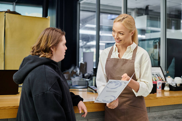 χαμογελαστός διευθυντής καφετέριας δείχνει κάρτα μενού σε νεαρή γυναίκα υπάλληλος με σύνδρομο down, inclusivity - Φωτογραφία, εικόνα