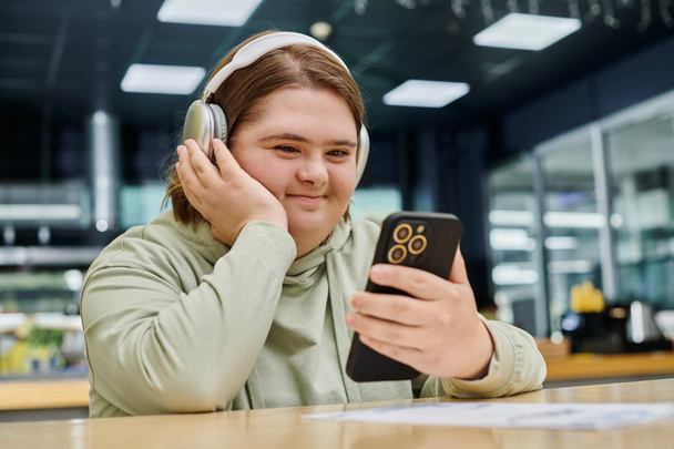 fröhliche Frau mit geistiger Störung hält Smartphone in der Hand und hört Musik über Kopfhörer im Café - Foto, Bild