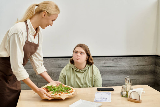 alegre camarera proponiendo deliciosa pizza a cliente femenino con síndrome de Down sentado en acogedora cafetería - Foto, imagen