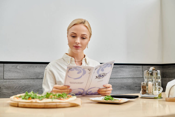mooie blonde vrouw op zoek in de menukaart in de buurt van heerlijke pizza tijdens het zitten in modern cafe - Foto, afbeelding