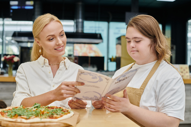 młoda kelnerka z zespołem Downa pokazująca kartę menu uśmiechniętej kobiecie w pobliżu pizzy w nowoczesnej kawiarni - Zdjęcie, obraz