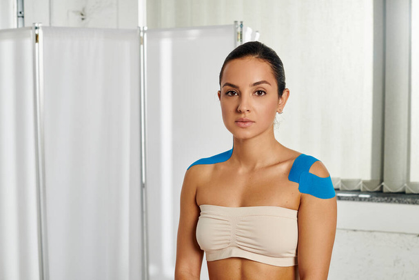 привлекательная молодая пациентка с кинезиологическими лентами на плечах, смотрящая в камеру, здравоохранение - Фото, изображение