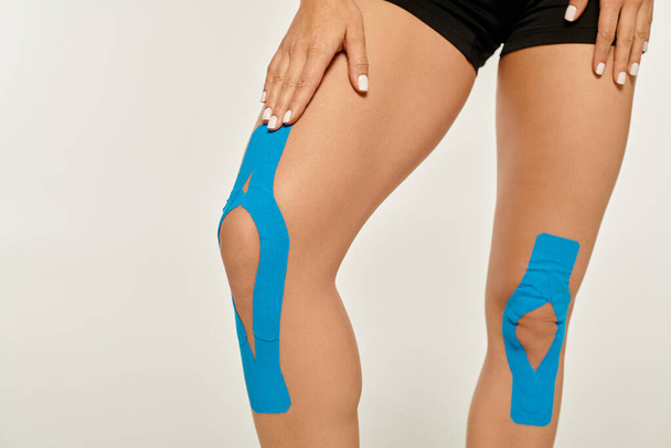 bijgesneden weergave van jonge vrouw in sport slijtage met kinesiologische tapes op haar knieën op grijze achtergrond - Foto, afbeelding