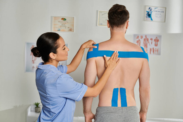 beautiful young doctor in medical costume putting kinesio tapes on shoulders of her bearded patient - Photo, Image