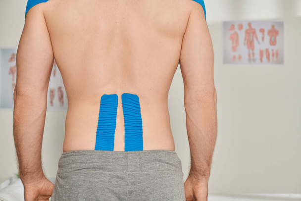 abgeschnittene Rückenansicht eines Mannes in grauer Jogginghose mit kinesiologischen Klebebändern auf dem Rücken, Gesundheitswesen - Foto, Bild