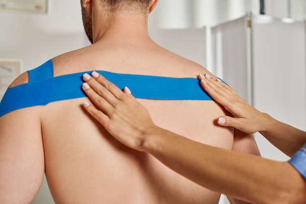 Ausgeschnittene Ansicht der Hände einer jungen Ärztin, die ihrem männlichen Patienten kinesiologisches Klebeband auf den Rücken legt - Foto, Bild