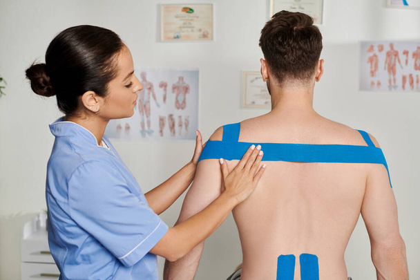 attraktive Ärztin in blauem Medizinkostüm, die Kinesiobänder auf den Rücken ihrer Patientin legt, Gesundheitswesen - Foto, Bild