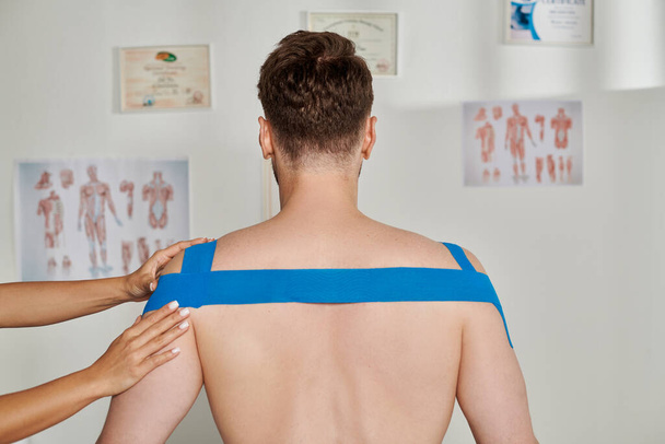 Omuzlarına ve sırtına kinezyolojik bantlar yapıştıran kadın eli olan bir adamın arka görüntüsü. - Fotoğraf, Görsel