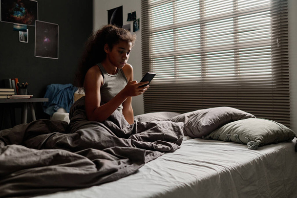 Linda adolescente afroamericana mirando a través de mensajes de acoso cibernético de usuarios anónimos mientras está sentada en la cama por la mañana - Foto, imagen