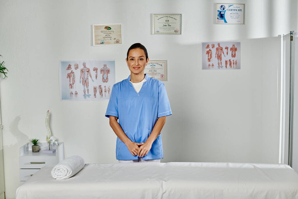 Χαρούμενη νεαρή γυναίκα γιατρός με ιατρική στολή ποζάρει στο γραφείο της και χαμογελάει στην κάμερα - Φωτογραφία, εικόνα