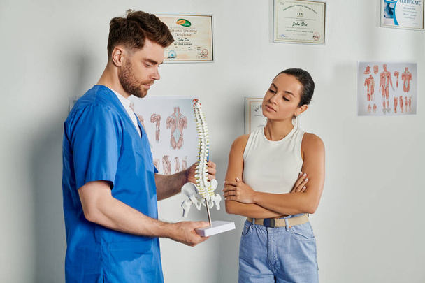 attraktive Patientin beim Anblick eines gutaussehenden bärtigen Arztes mit Wirbelsäulenmodell in seinen Händen - Foto, Bild