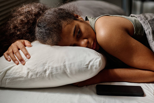 Депресивна дівчина-підліток тримає голову на подушці, лежачи в ліжку і дивлячись на смартфон з повідомленнями про кіберзалякування від анонімного користувача - Фото, зображення