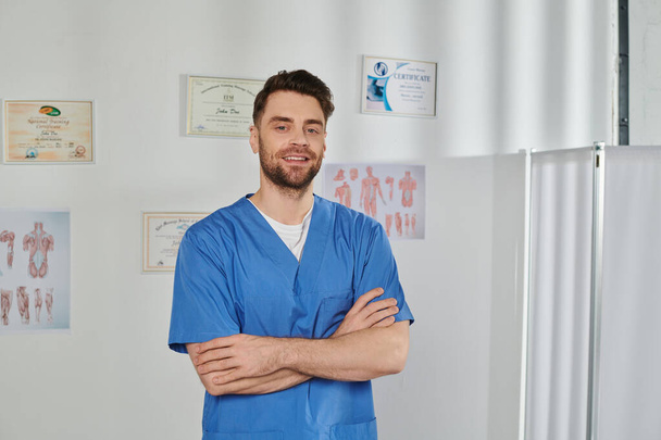 alegre médico guapo posando con los brazos cruzados en el pecho y sonriendo a la cámara, atención médica - Foto, imagen