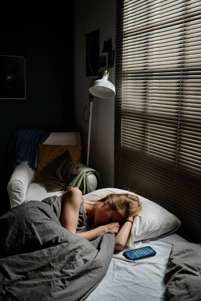 Дівчинка-підліток лежить на ліжку біля вікна, вкрита венеціанськими жалюзями рано вранці і плаче, страждаючи від кіберзалякування - Фото, зображення