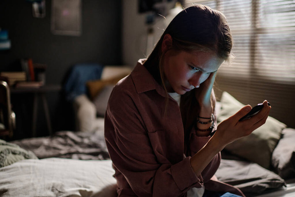 Obrażona nastolatka ze smartfonem cierpiąca na cybernękanie w dzień i w nocy podczas przeglądania anonimowych wiadomości - Zdjęcie, obraz