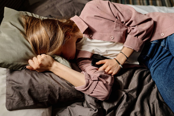 Gefrustreerde tiener met smartphone in de hand huilen in kussen vanwege cyberpesten berichten van anonieme mensen terwijl ze in bed liggen - Foto, afbeelding